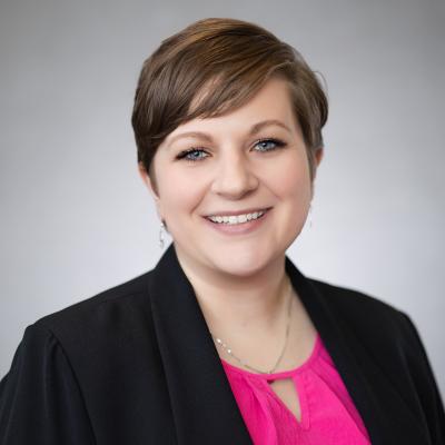 A light complexion female with brown hair and blue eyes smiles at the camera and wears a black blazer, a hot pink shirt and a silver necklace.