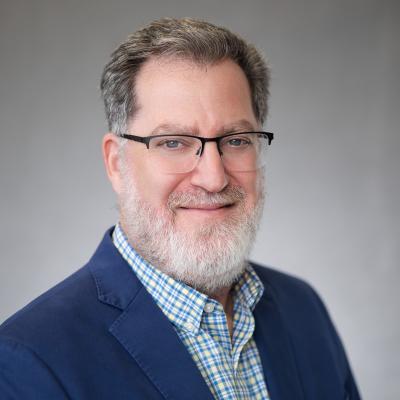 A light complexion male with blue eyes and grey hair smiles at the camera and is wearing a navy blue blazer with a button up shirt with a checkered pattern.