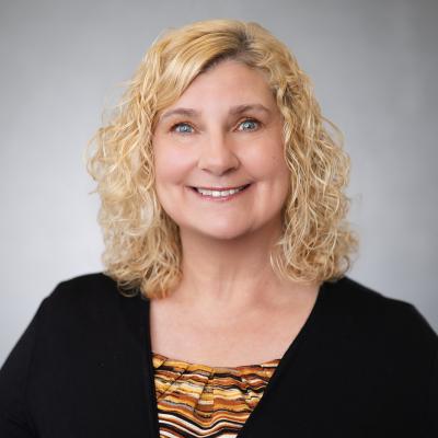 A light complexion female with blonde hair and green eyes smiles at the camera and wears a black blazer with an orange, yellow black and white patterned shirt.