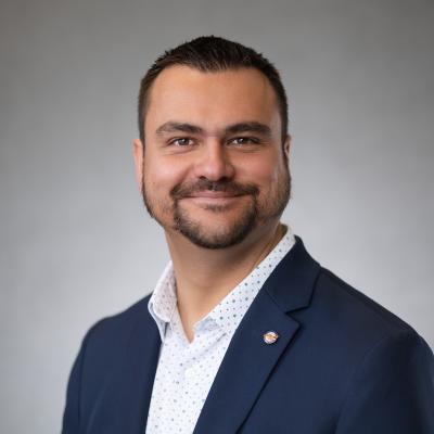 A tan complexioned male  with brown eyes and black ahir smiles at the camera and wears a navy blue blazer with a dotted white button up shirt.