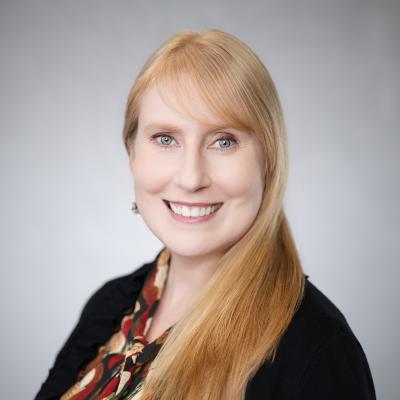 A light complexion female with blue eyes and strawberry blonde hair smiles at the camera and is wearing a black blazer with a red, orange and black patterned shirt.