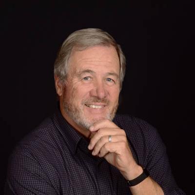 Gerry Bird sits and smiles with a hand touching his chin. He is wearing a blue shirt against a black backgorund with a black bracelet on his wrist.