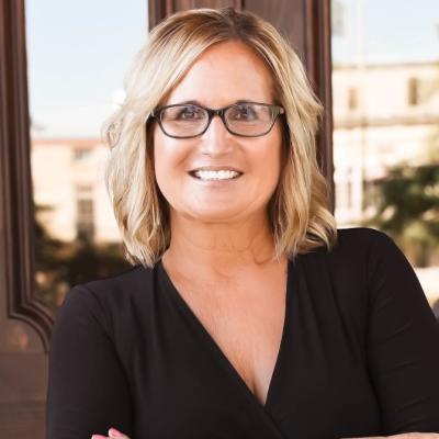 Jennifer McDonald stands in front of an open window with a courthouse-like building in the background. She is blonde, wearing glasses and wearing a black shirt.
