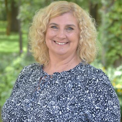 A light complexion female with blonde hair wearing a blue blouse with a white pattern smiles at the viewer.