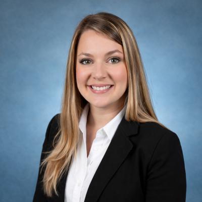 This is a photo of a light-skinned woman with long, blond hair, smiling, wearing a black blazer and a white button-up shirt against a blue backgroud.