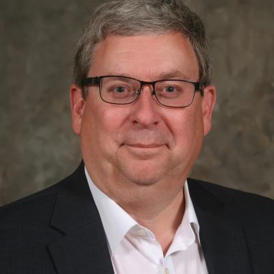Rick Platt sits and smiles with no background behind him except a grey screen. He is wearing a blue blazer and a white button-down shirt. He is wearing black glasses. 